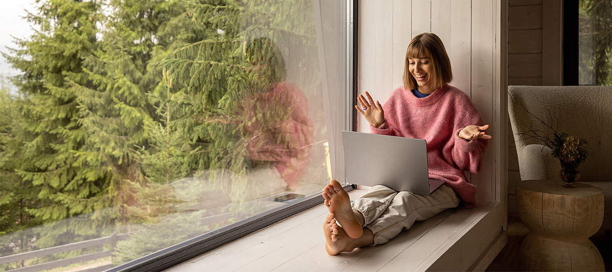 junge Frau sitzt gemütlich auf breiter Fensterbank an großem Fenster mit Sicht auf Wald und freut sich über etwas, das sie im Laptop sieht